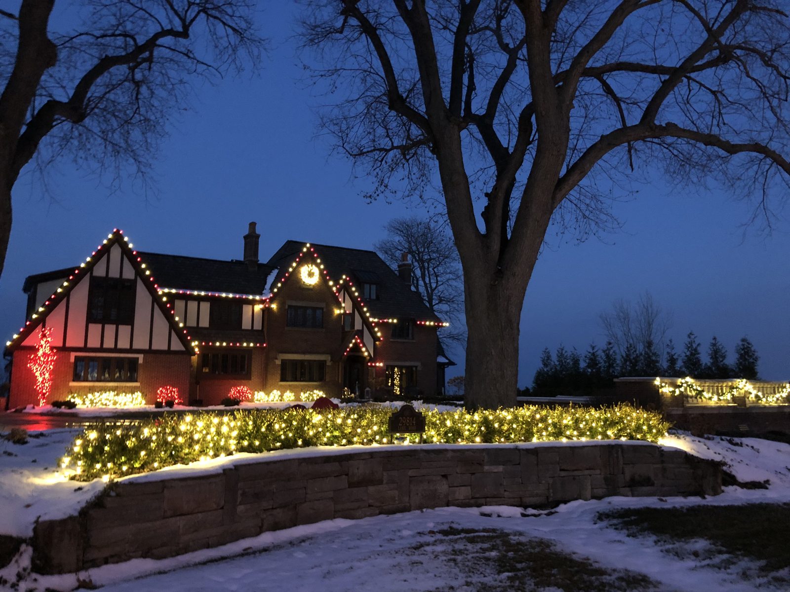 Tudor-style Cleveland-area home with professional Christmas lights along the rooflines, a lit wreath, and warm white lights on hedges and shrubs in the snow